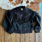 Black fringed jacket on a wooden surface with a cowhide rug in the background
