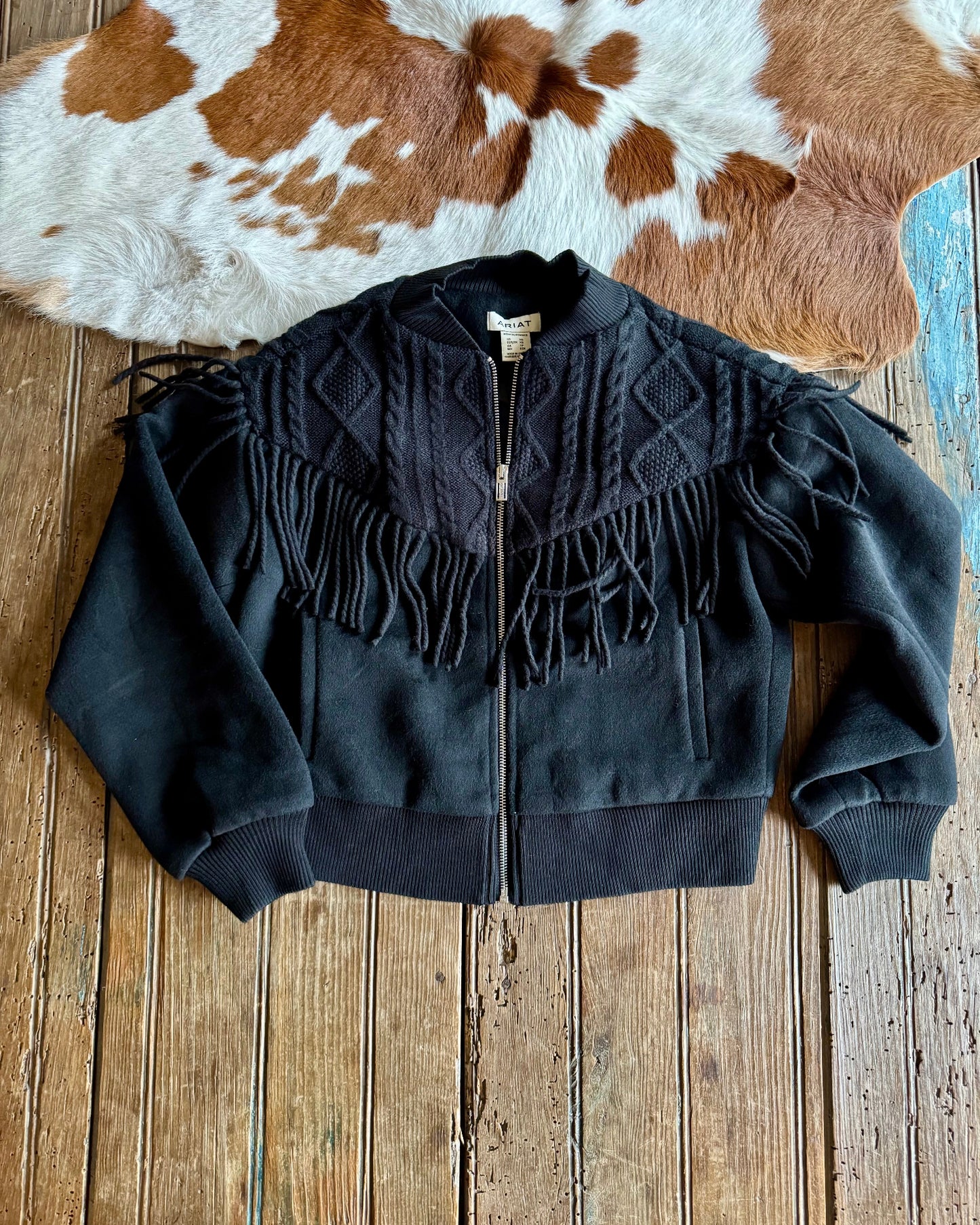 Black fringed jacket on a wooden surface with a cowhide rug in the background