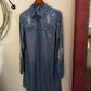 Blue denim shirt with embroidered designs on a hanger against a white wall.