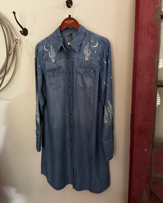 Blue denim shirt with embroidered designs on a hanger against a white wall.
