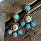 A pair of silver-toned dangle earrings with turquoise-blue stones and white inclusions, displayed on a rustic wooden surface next to a metal ruler for scale.