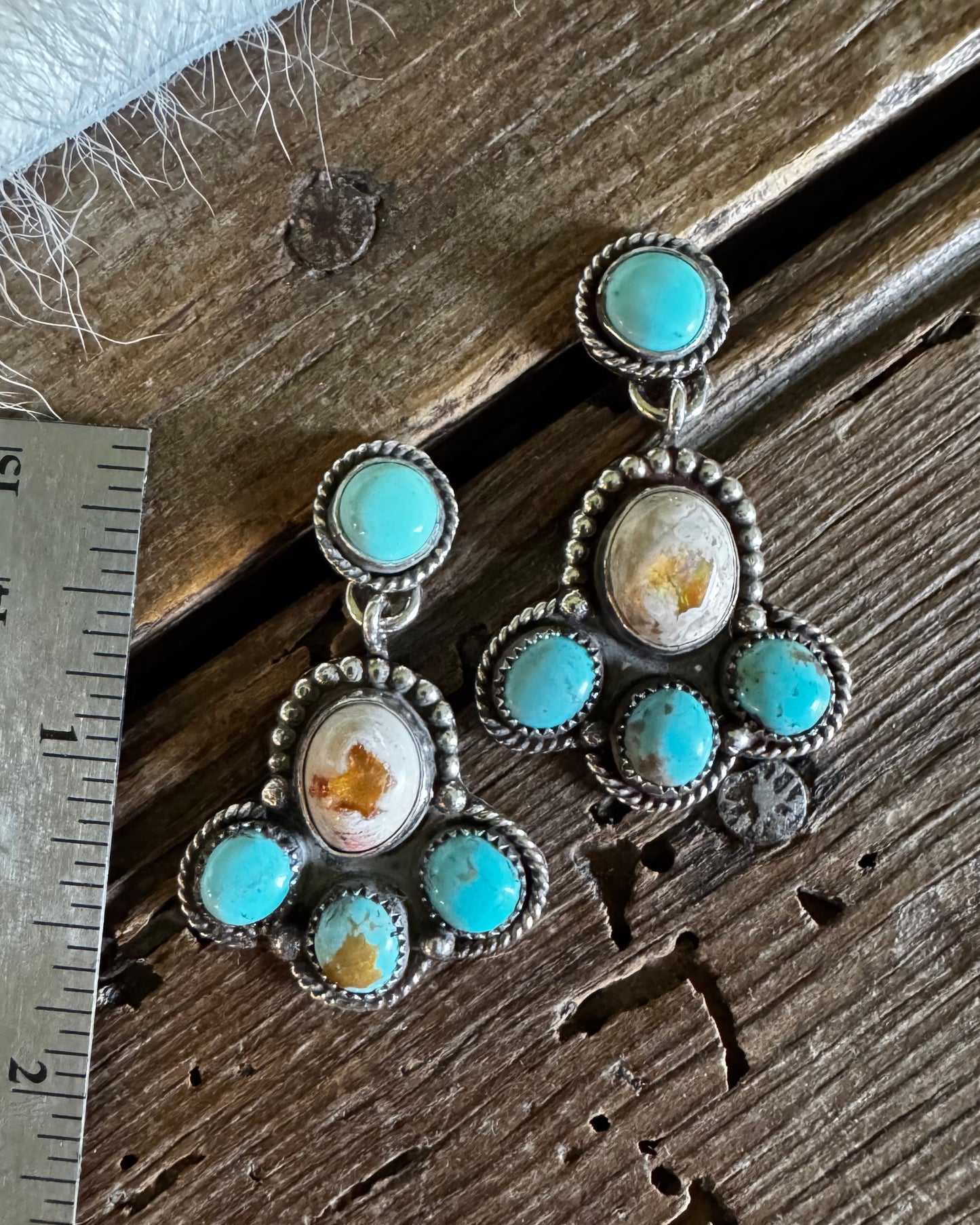 A pair of silver-toned dangle earrings with turquoise-blue stones and white inclusions, displayed on a rustic wooden surface next to a metal ruler for scale.