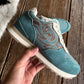 A teal suede sneaker with brown embroidered swirl patterns and cream laces, held by a hand over a rustic wooden plank floor.