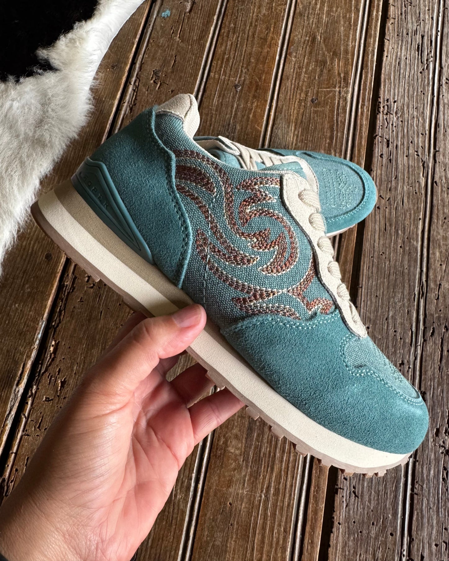 A teal suede sneaker with brown embroidered swirl patterns and cream laces, held by a hand over a rustic wooden plank floor.