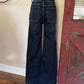Wide-leg dark blue denim jeans shown from the back on a black mannequin torso, standing on a rolling display stand in an indoor room with a beige wall, wood floor, and distressed red door.