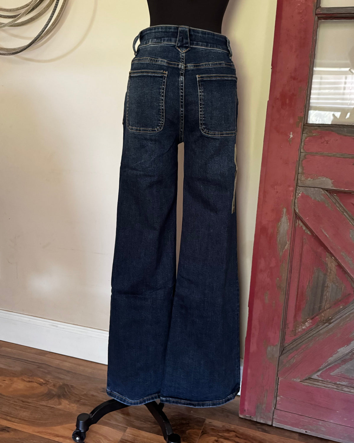 Wide-leg dark blue denim jeans shown from the back on a black mannequin torso, standing on a rolling display stand in an indoor room with a beige wall, wood floor, and distressed red door.