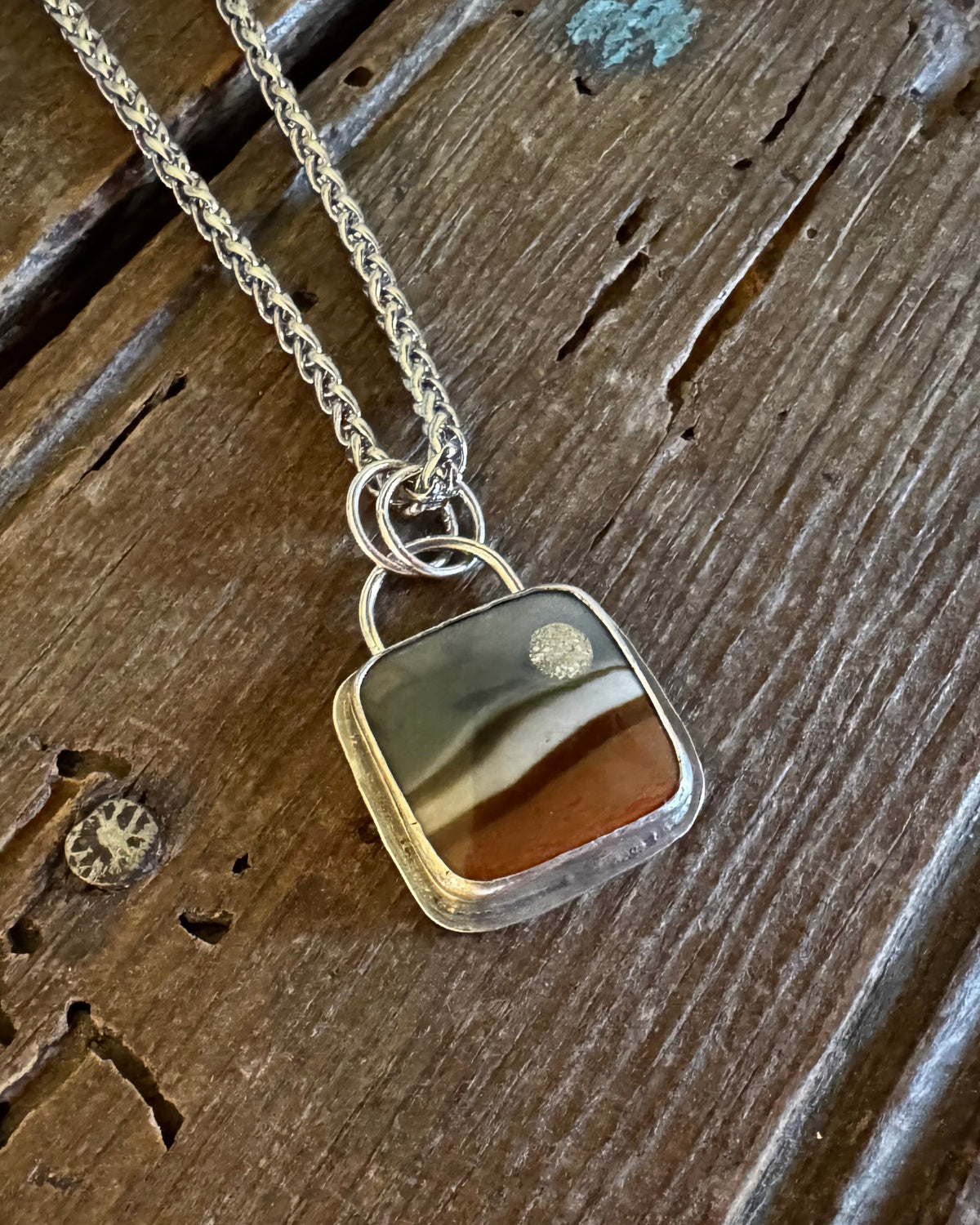 A silver-toned chain necklace with a square pendant featuring a marbled pattern of brown, beige, and gray, resting on a rustic dark wood surface.
