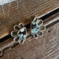 A pair of silver-toned stud earrings with a floral or fan-shaped outline and a central triangular turquoise stone, displayed on a rustic wooden surface with white faux fur.