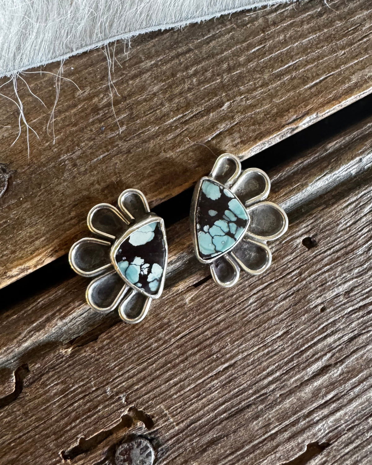 A pair of silver-toned stud earrings with a floral or fan-shaped outline and a central triangular turquoise stone, displayed on a rustic wooden surface with white faux fur.