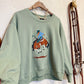 A sage green crewneck sweatshirt hanging on a wooden hanger, featuring a graphic of a cowboy riding a bucking horse on the front. The sweatshirt is styled with a gold chain necklace, set against a textured white wall with rustic western-themed decor on the right.