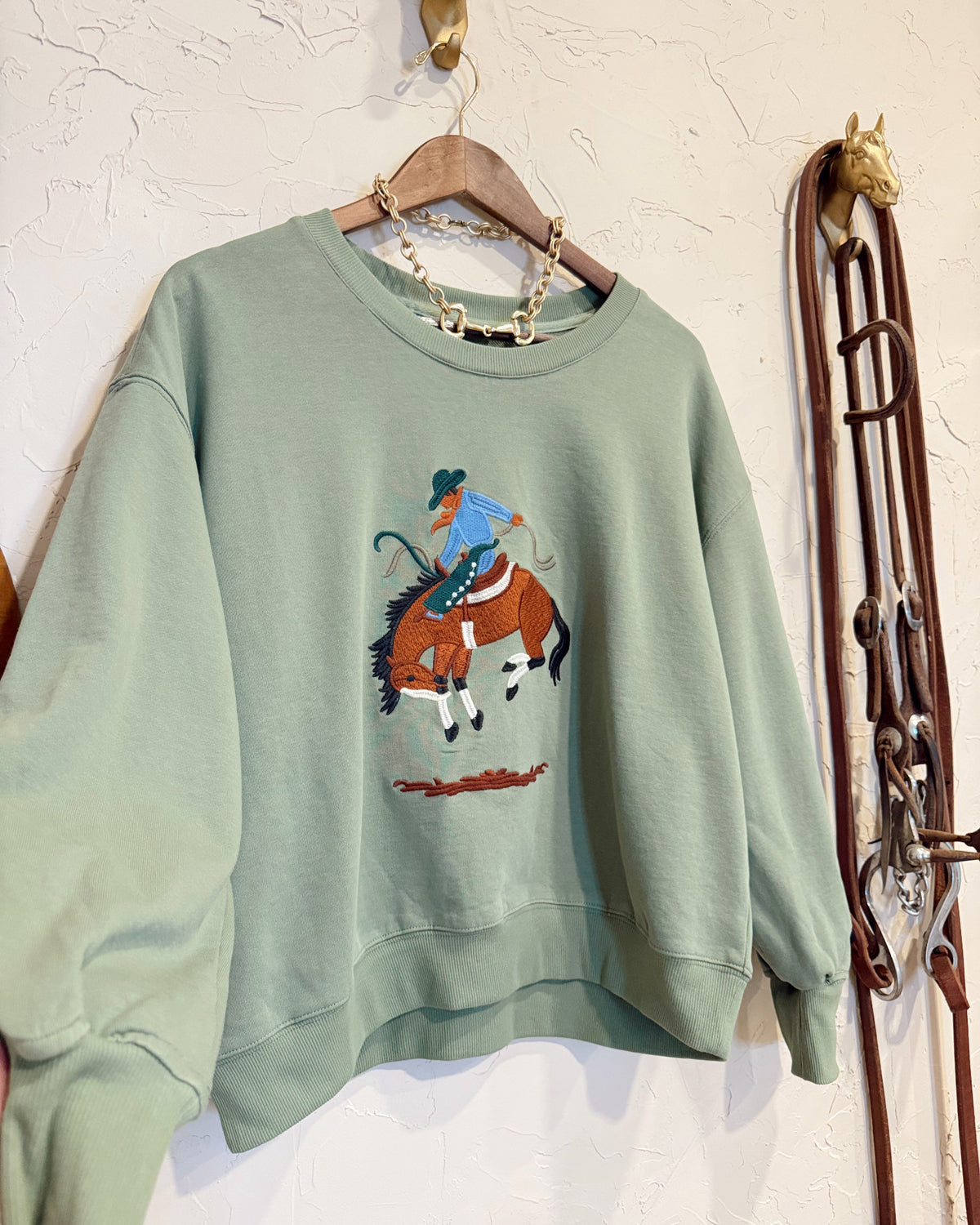 A sage green crewneck sweatshirt hanging on a wooden hanger, featuring a graphic of a cowboy riding a bucking horse on the front. The sweatshirt is styled with a gold chain necklace, set against a textured white wall with rustic western-themed decor on the right.