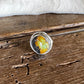 A silver-toned ring with a round cabochon containing yellow, orange, and green inclusions, resting on a rustic dark wood surface with a white faux-fur trim in the background.