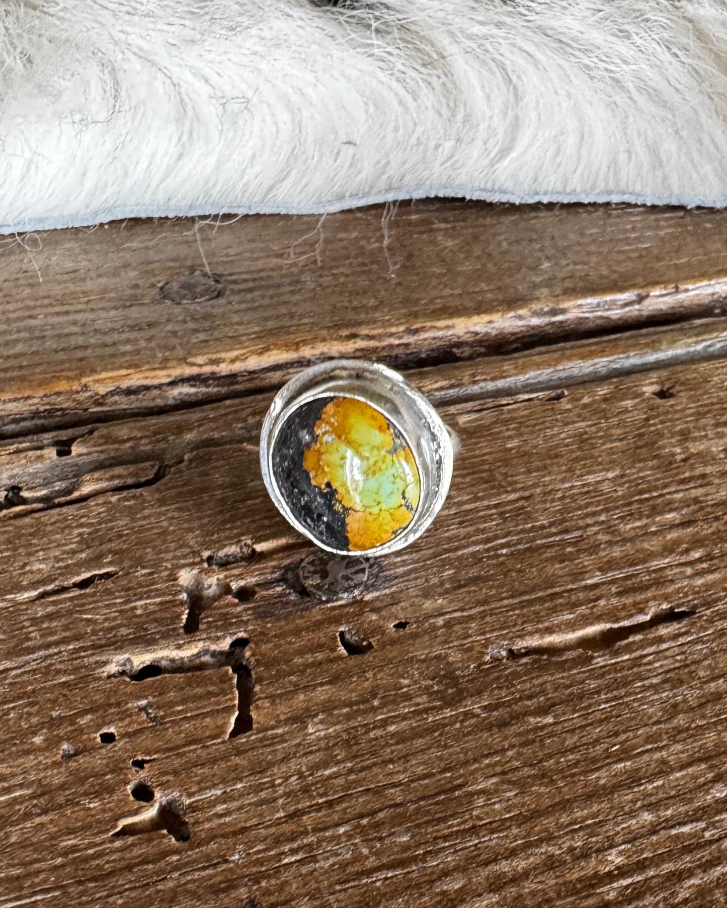 A silver-toned ring with a round cabochon containing yellow, orange, and green inclusions, resting on a rustic dark wood surface with a white faux-fur trim in the background.