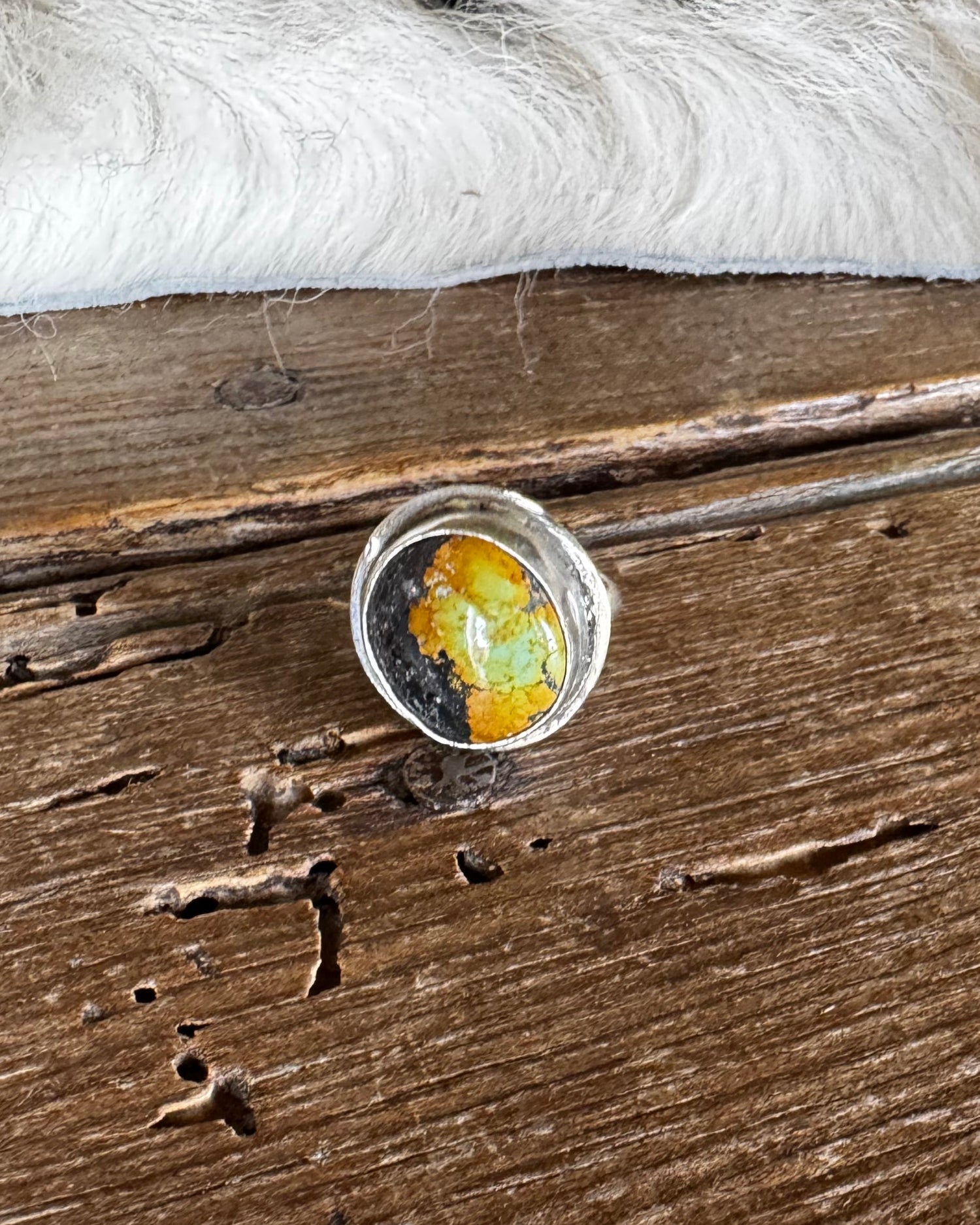 A silver-toned ring with a round cabochon containing yellow, orange, and green inclusions, resting on a rustic dark wood surface with a white faux-fur trim in the background.