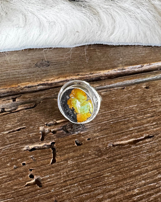A silver-toned ring with a round cabochon containing yellow, orange, and green inclusions, resting on a rustic dark wood surface with a white faux-fur trim in the background.