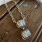 A handmade sterling silver necklace with two square turquoise and agate pendants, displayed on a rustic dark wood surface with a white faux fur accent.