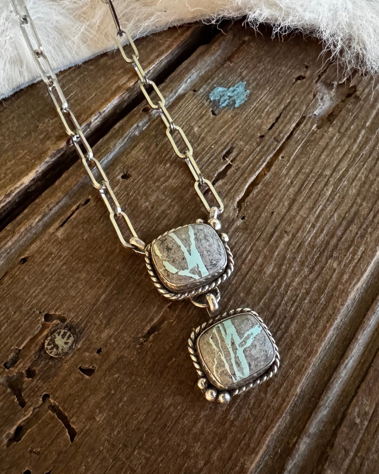 A handmade sterling silver necklace with two square turquoise and agate pendants, displayed on a rustic dark wood surface with a white faux fur accent.