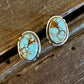A pair of oval stud earrings with turquoise-blue marbled stones and gold-toned settings, resting on a rustic wooden surface with a white faux-fur texture above.