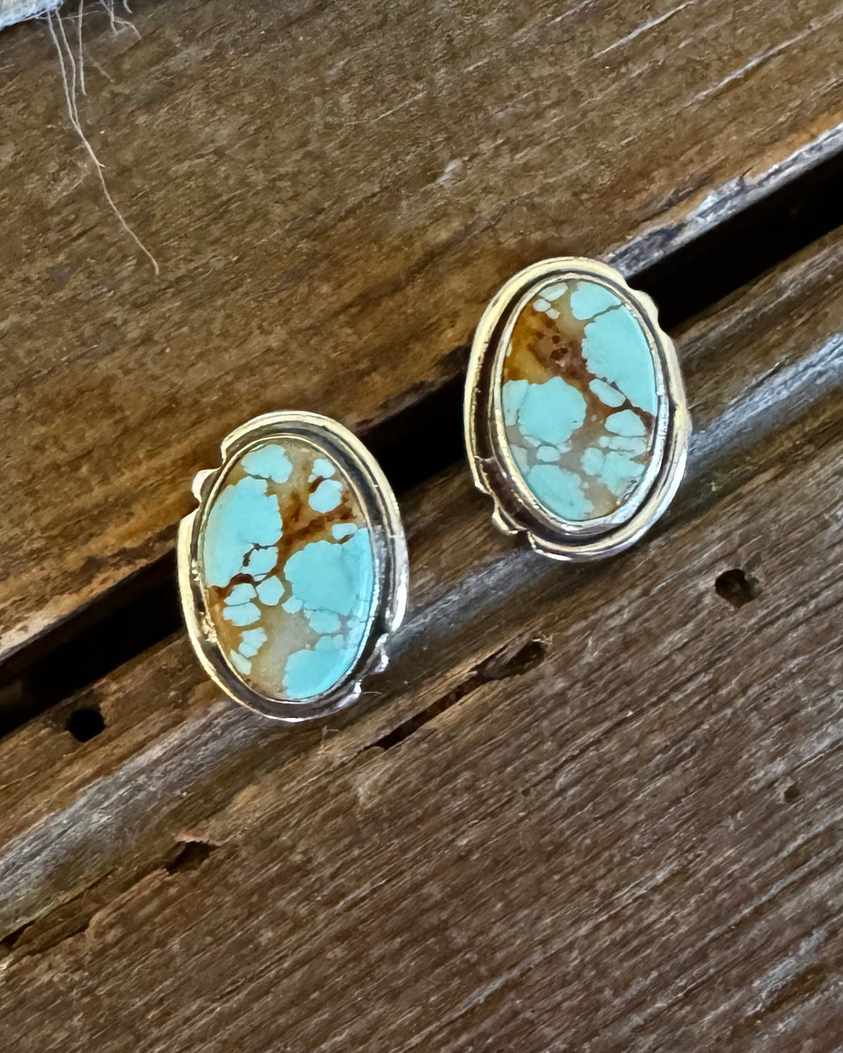A pair of oval stud earrings with turquoise-blue marbled stones and gold-toned settings, resting on a rustic wooden surface with a white faux-fur texture above.