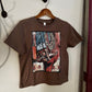 A brown short-sleeve t-shirt hanging on a wooden hanger against a white wall. The shirt features a large, colorful graphic print on the front depicting a cowboy boot and western elements in a cubist style.