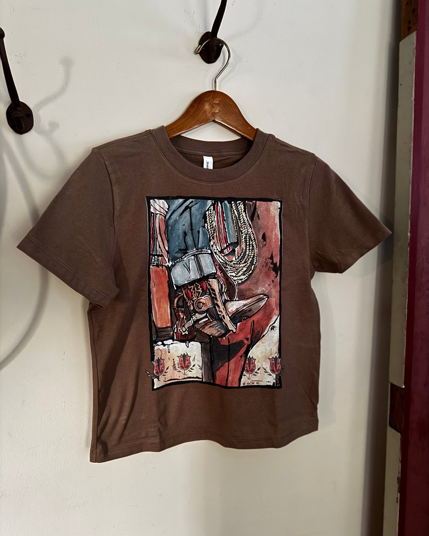 A brown short-sleeve t-shirt hanging on a wooden hanger against a white wall. The shirt features a large, colorful graphic print on the front depicting a cowboy boot and western elements in a cubist style.