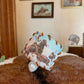 A small square fabric tag or patch with a western cowboy and horse print, attached to a white plastic clip, resting on a brown and white cowhide rug in a room with framed wall art.