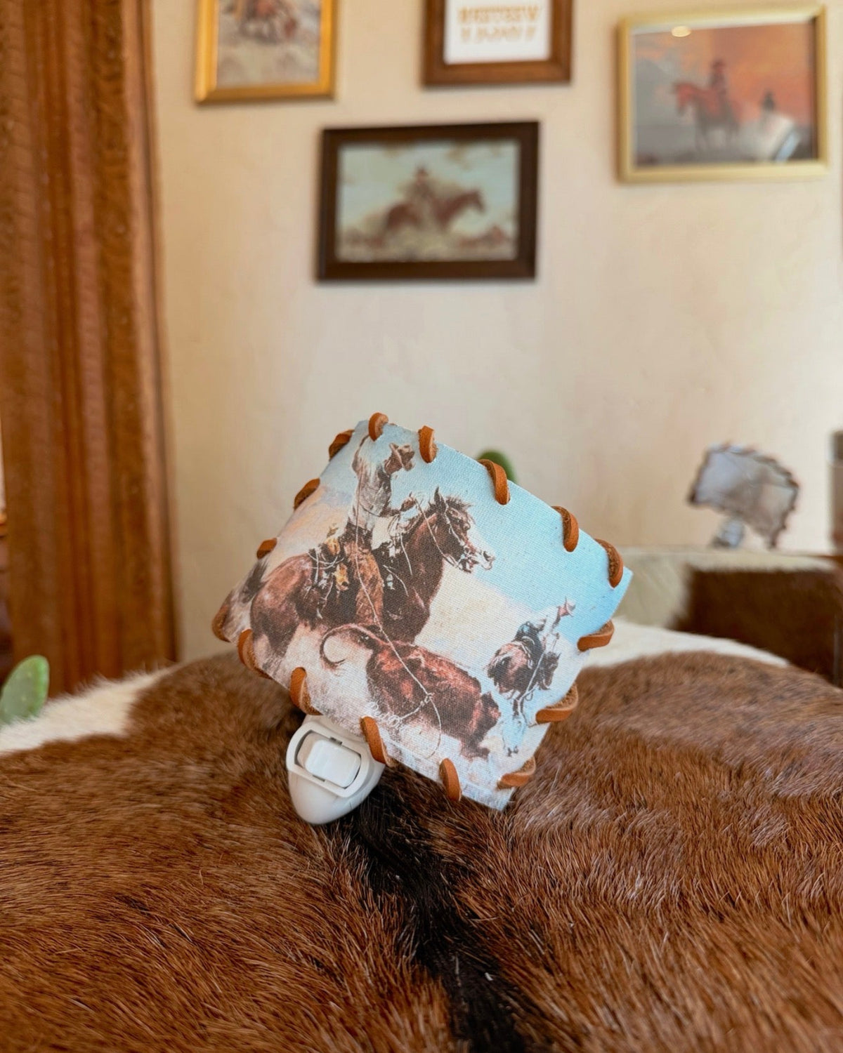 A small square fabric tag or patch with a western cowboy and horse print, attached to a white plastic clip, resting on a brown and white cowhide rug in a room with framed wall art.