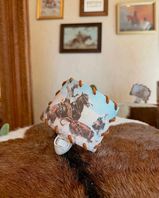 A small square fabric tag or patch with a western cowboy and horse print, attached to a white plastic clip, resting on a brown and white cowhide rug in a room with framed wall art.