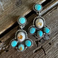 A pair of silver-toned dangle earrings with turquoise and natural stone accents, displayed on a rustic wooden surface.