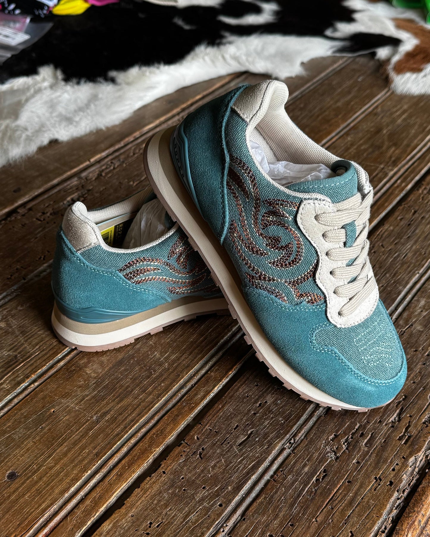 A pair of turquoise western-style sneakers with white laces and brown embroidered details, resting on a rustic wooden table in a cluttered indoor setting.