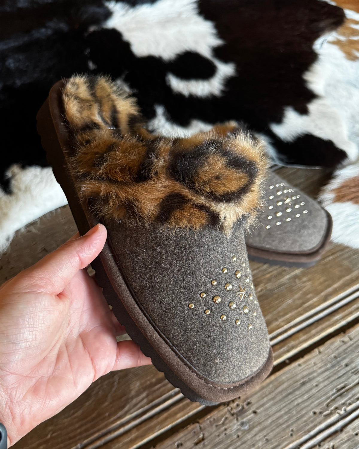 A hand holds a brown suede mule slipper with a leopard-print faux fur trim, set against a rustic wood floor and a black-and-white cowhide rug.