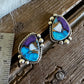 A pair of Southwestern-style turquoise and purple stone earrings with silver bezel settings, displayed on a rustic wooden surface next to a metal ruler for scale.
