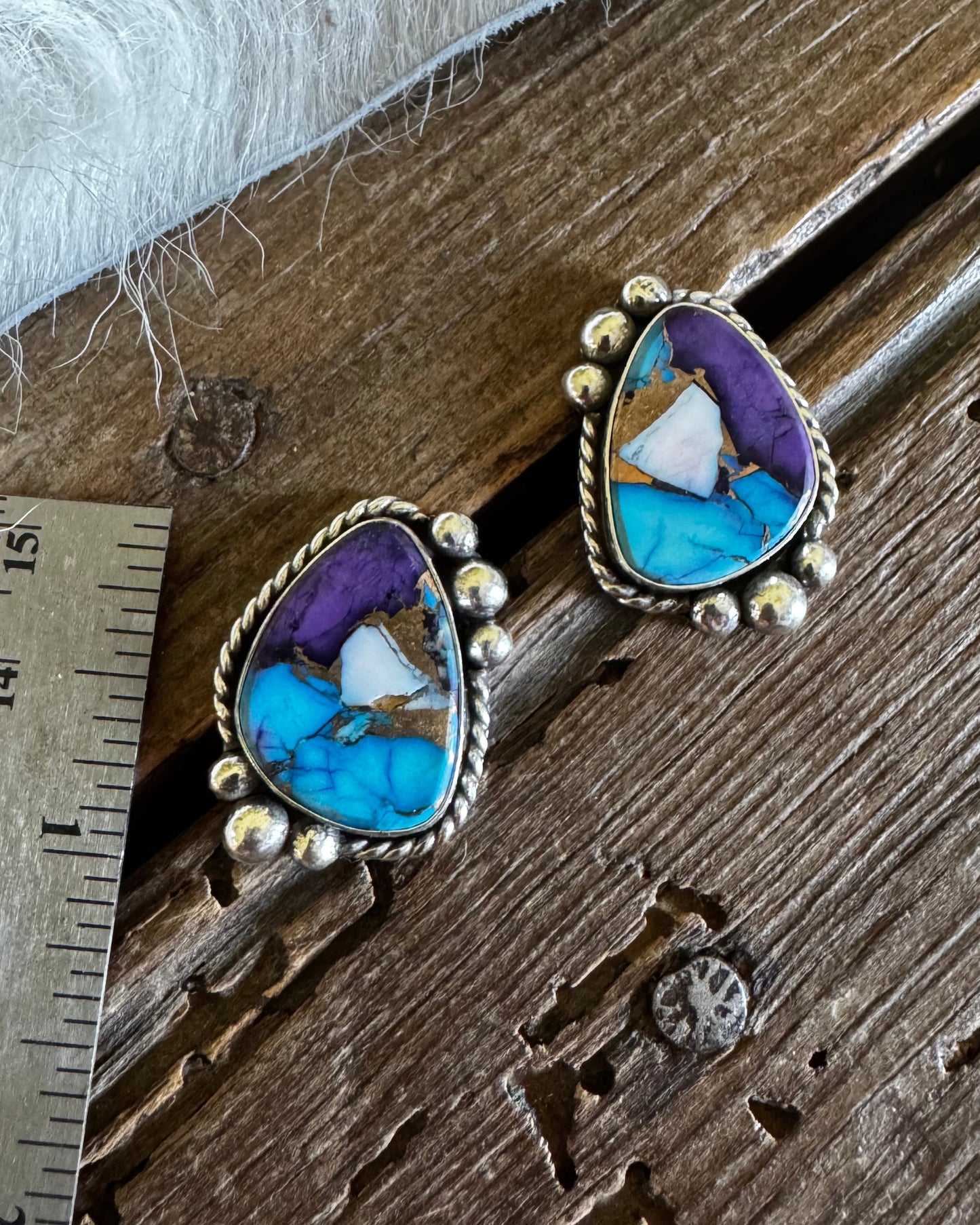 A pair of Southwestern-style turquoise and purple stone earrings with silver bezel settings, displayed on a rustic wooden surface next to a metal ruler for scale.
