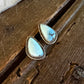 A pair of teardrop-shaped turquoise stud earrings with gold-toned bezel settings, resting on a rustic dark wood surface next to a white fur trim.