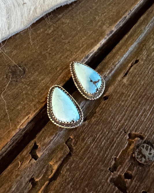 A pair of teardrop-shaped turquoise stud earrings with gold-toned bezel settings, resting on a rustic dark wood surface next to a white fur trim.