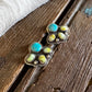 A pair of silver-toned stud earrings with turquoise and yellow-green stone accents, displayed on a rustic wooden surface with a white faux-fur trim at the top.