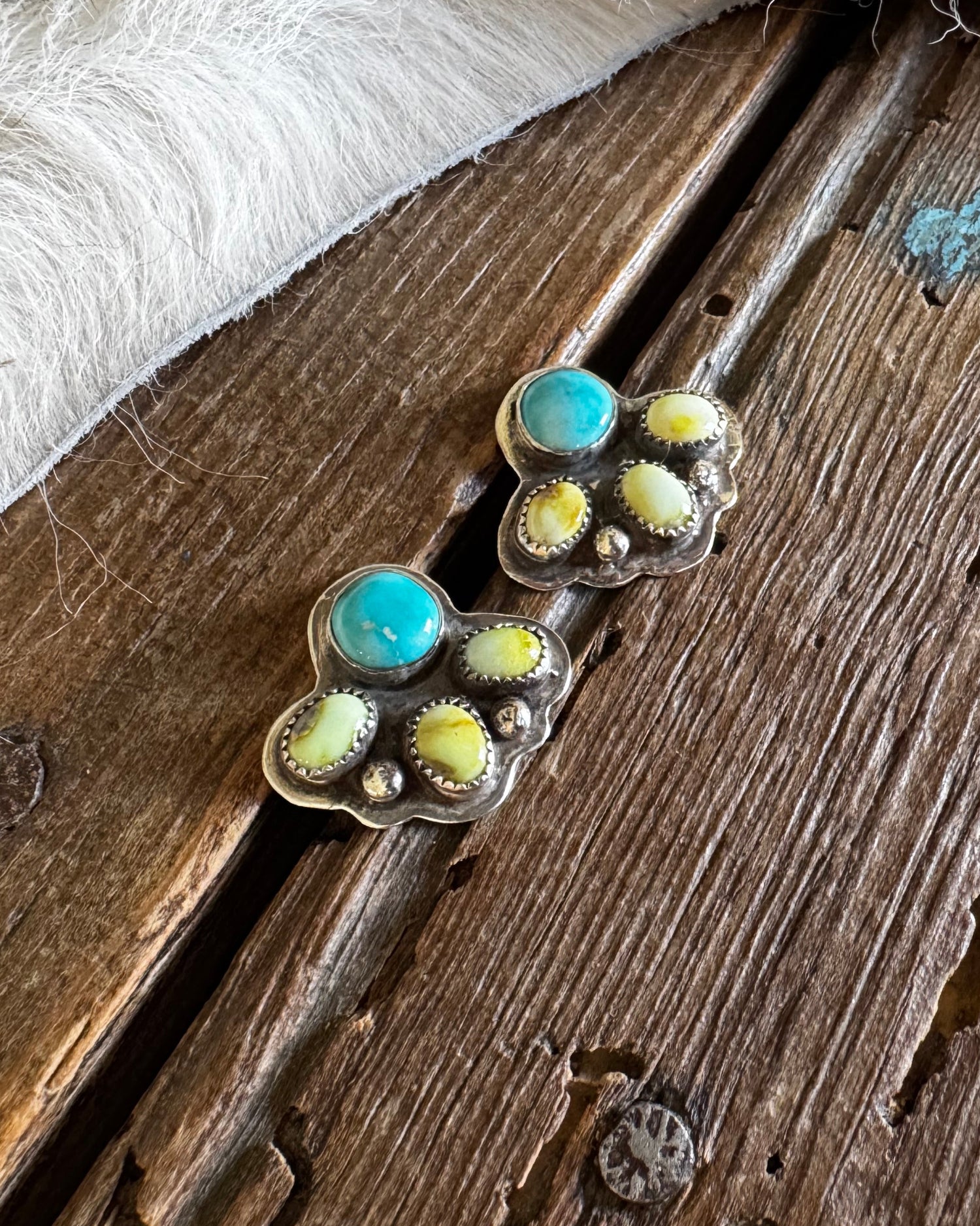 A pair of silver-toned stud earrings with turquoise and yellow-green stone accents, displayed on a rustic wooden surface with a white faux-fur trim at the top.