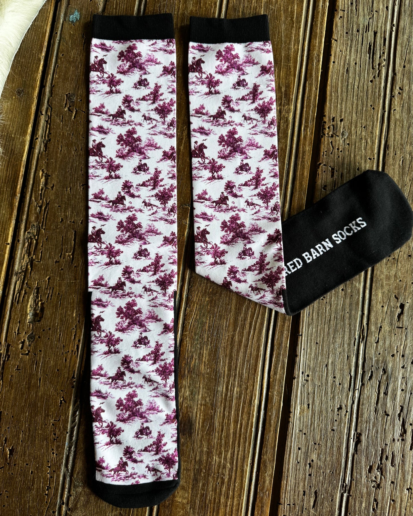 A pair of patterned knee-high socks and a black fabric drawstring bag on a rustic wooden surface. The socks have a white background with a dusty purple floral toile pattern and black cuffs and toes.