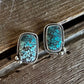 A pair of rectangular stud earrings with turquoise-blue and green marbled stone in a silver-toned bezel setting, resting on a rustic wooden surface.
