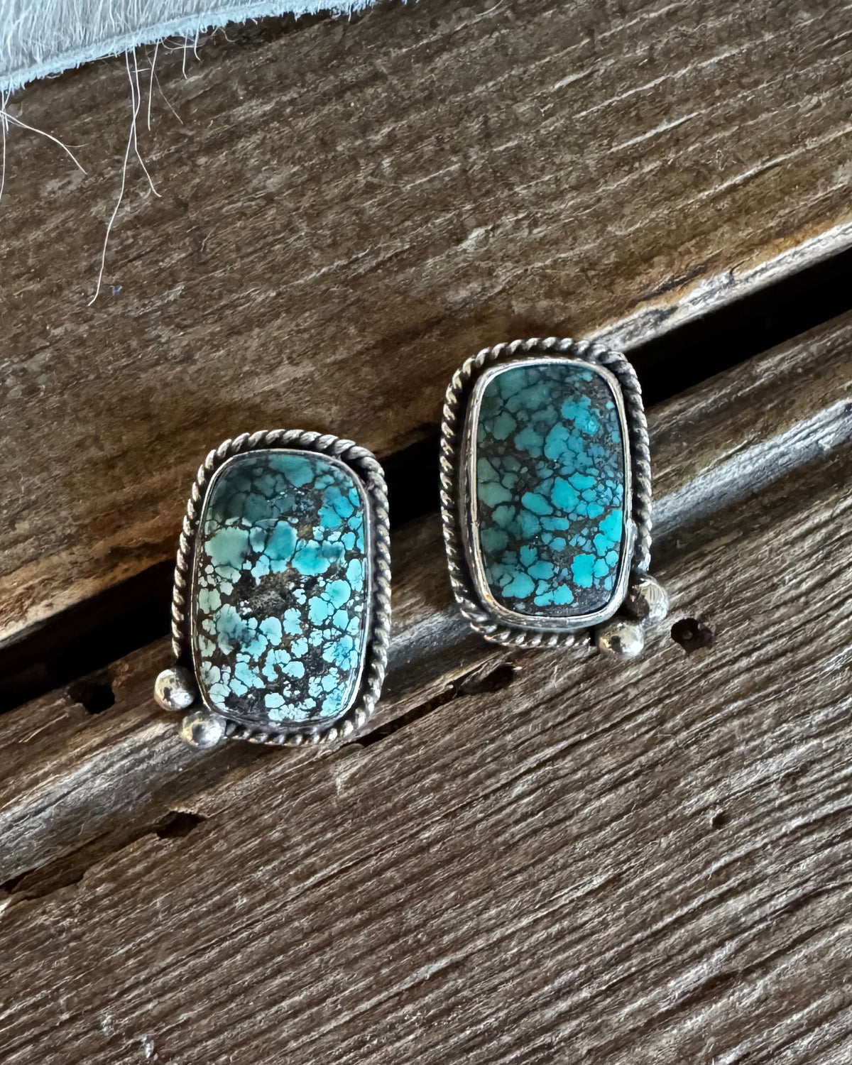A pair of rectangular stud earrings with turquoise-blue and green marbled stone in a silver-toned bezel setting, resting on a rustic wooden surface.