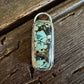 A rectangular silver-toned pendant featuring a natural turquoise stone with green, blue, and brown matrix patterns, resting on a rustic dark wood surface.