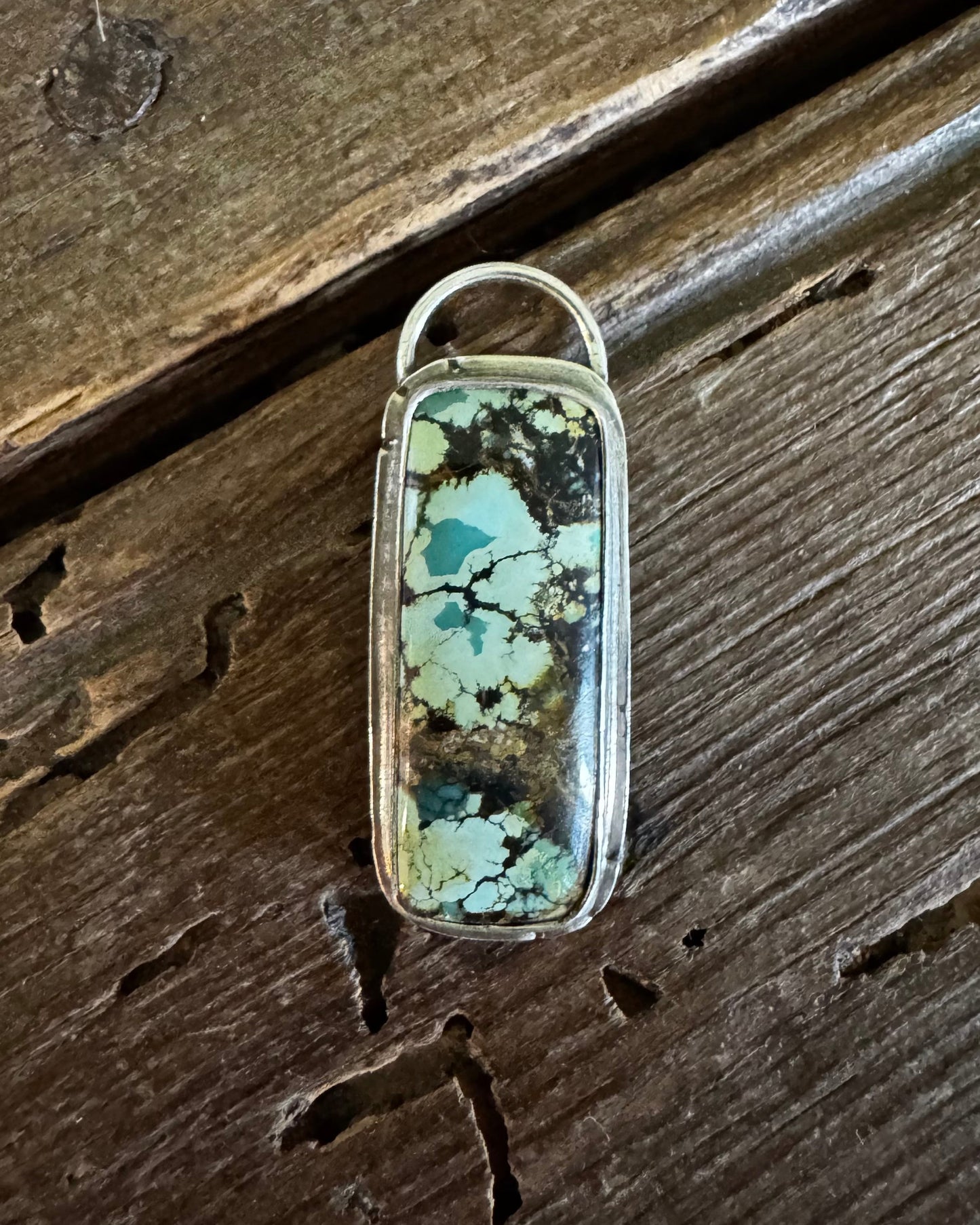 A rectangular silver-toned pendant featuring a natural turquoise stone with green, blue, and brown matrix patterns, resting on a rustic dark wood surface.