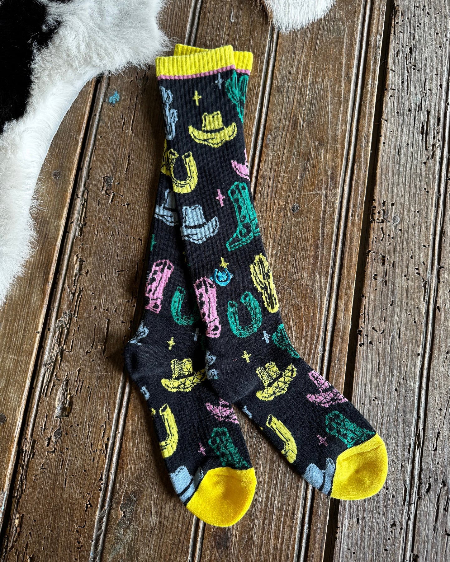 A pair of crew socks with a black background and a colorful western-themed print featuring yellow boots, pink horseshoes, green cacti, and blue hats, displayed on a rustic wooden surface.