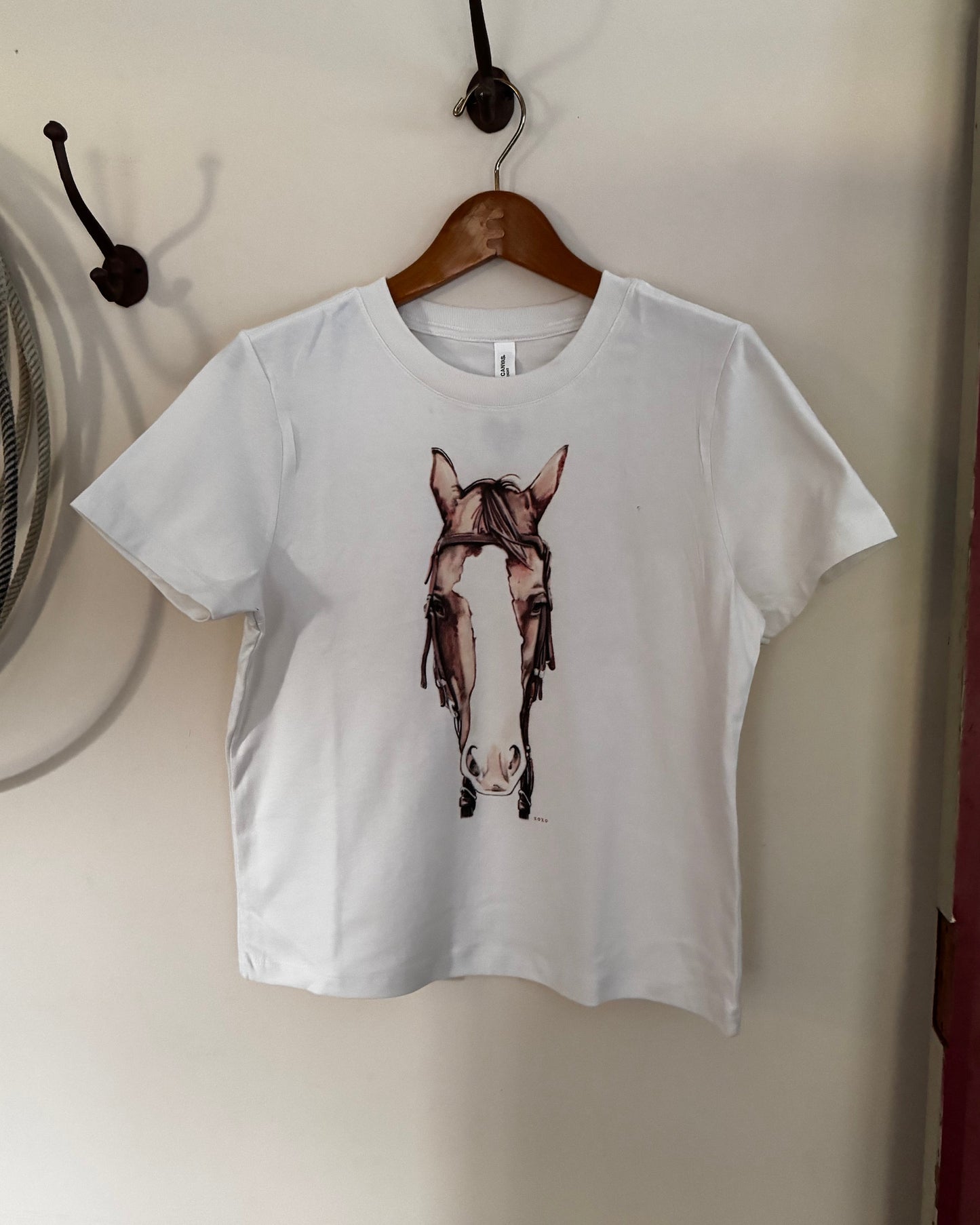 A white short-sleeve crop top t-shirt hanging on a wooden hanger from a black wall hook. The shirt features a graphic print of a horse's head wearing a bridle.