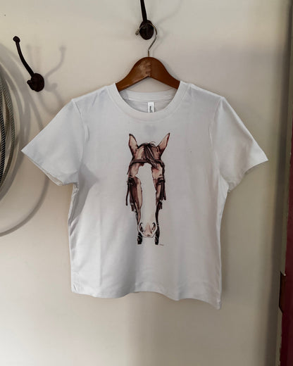 A white short-sleeve crop top t-shirt hanging on a wooden hanger from a black wall hook. The shirt features a graphic print of a horse's head wearing a bridle.