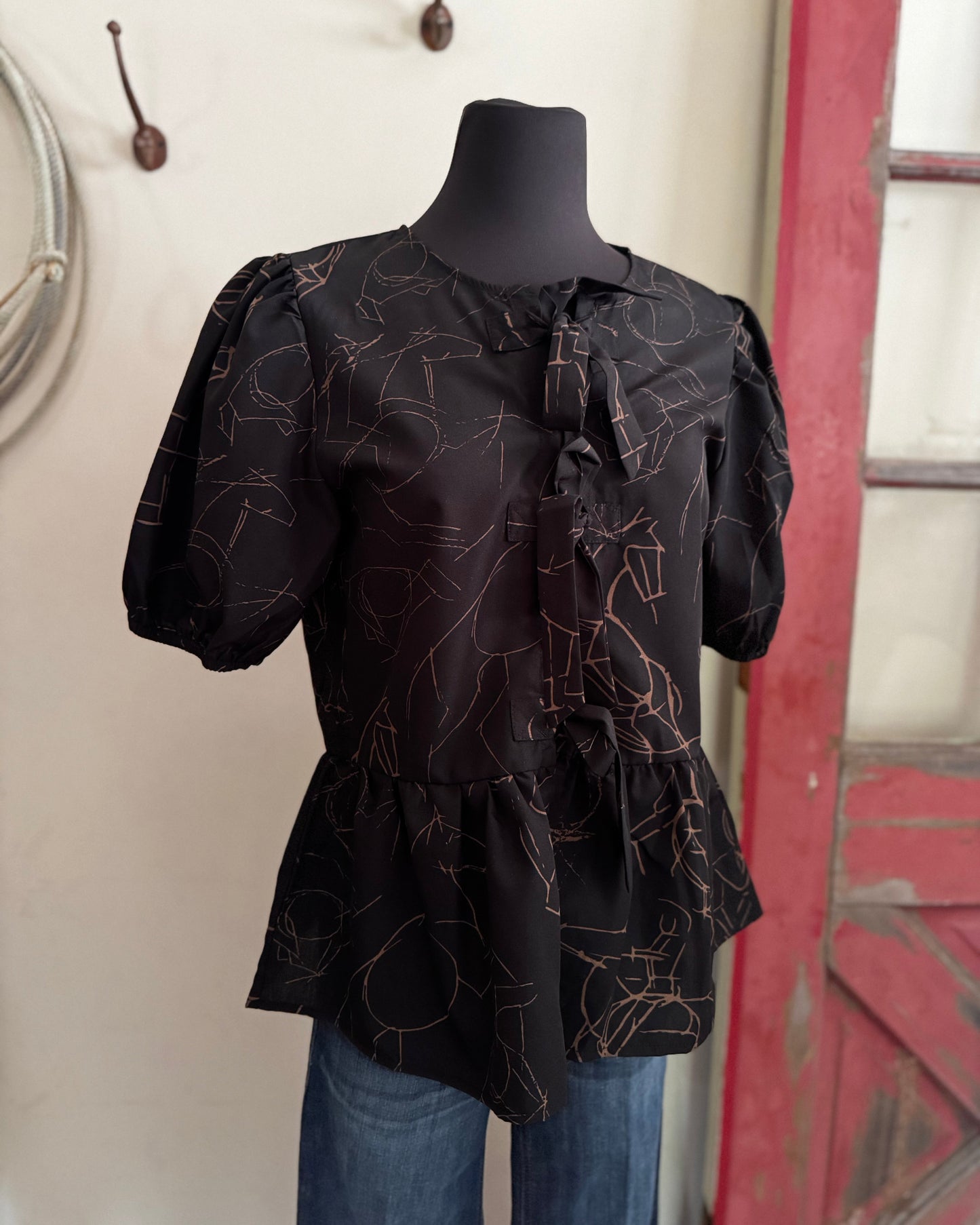 Black blouse with floral pattern on a mannequin against a neutral wall.