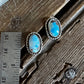 A pair of silver-toned oval stud earrings with bright blue opal-like centers, placed on a rustic wooden surface next to a metal ruler for scale and a white fur trim.