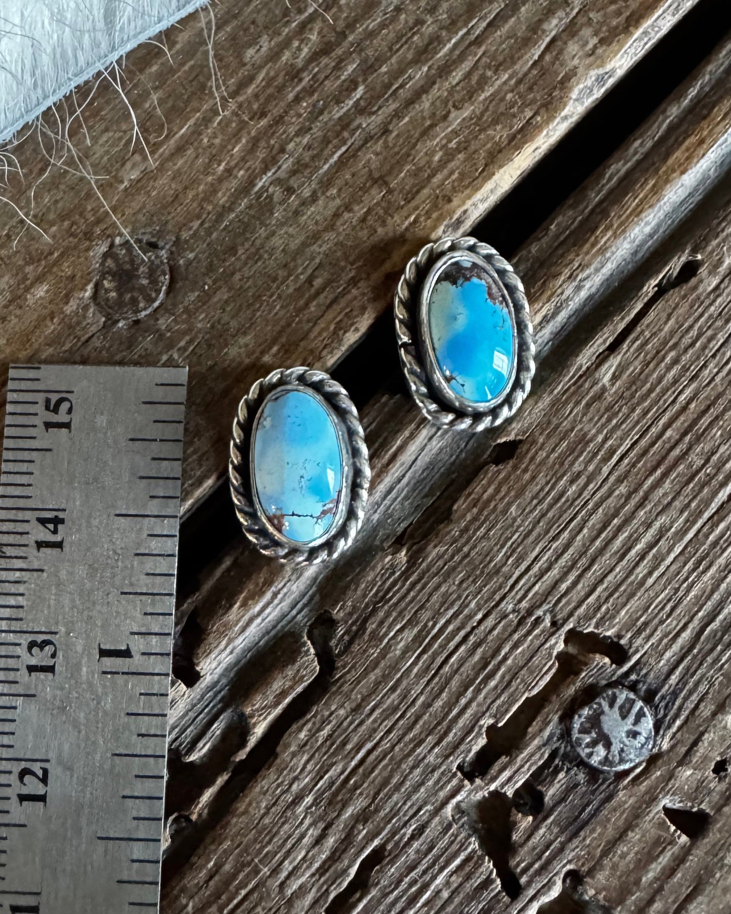 A pair of silver-toned oval stud earrings with bright blue opal-like centers, placed on a rustic wooden surface next to a metal ruler for scale and a white fur trim.