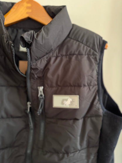 Black puffer vest with a brand logo on a hanger against a beige wall.
