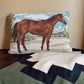 A rectangular needlepoint pillow featuring a brown horse in a field under a blue sky, displayed on a dark wooden surface against a beige wall, with a green and white chevron blanket in the foreground.